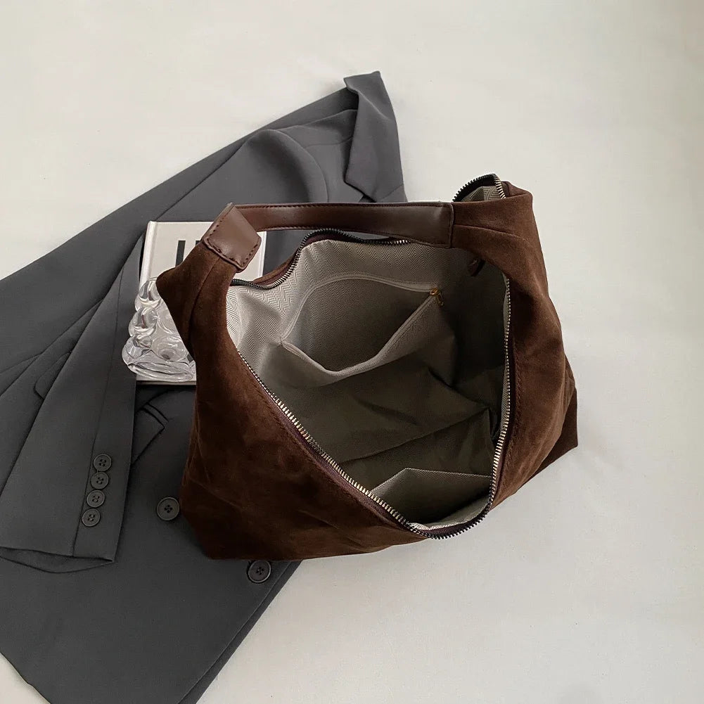 A brown suede slouchy shoulder bag is shown next to a gray blazer on a white background.