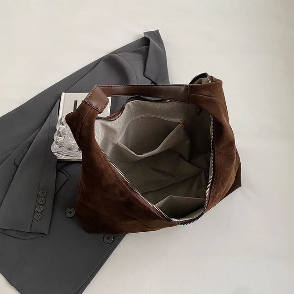 A brown suede slouchy shoulder bag is shown next to a gray blazer on a white background.