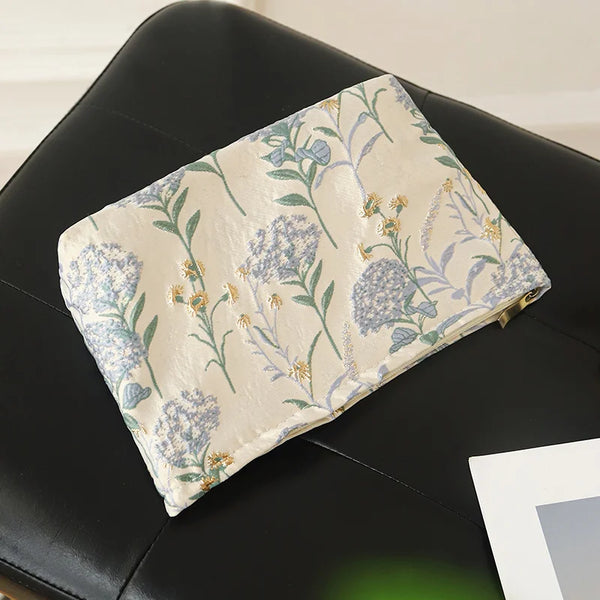 A floral canvas makeup bag featuring blue and green botanical embroidery resting on a black surface.