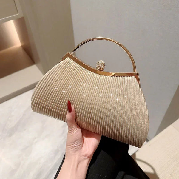 A beige pleated evening handbag with a gold top handle and chain strap is held by a hand.