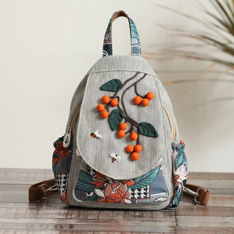 A handcrafted linen backpack featuring green leaves and orange berries embroidered on the front.