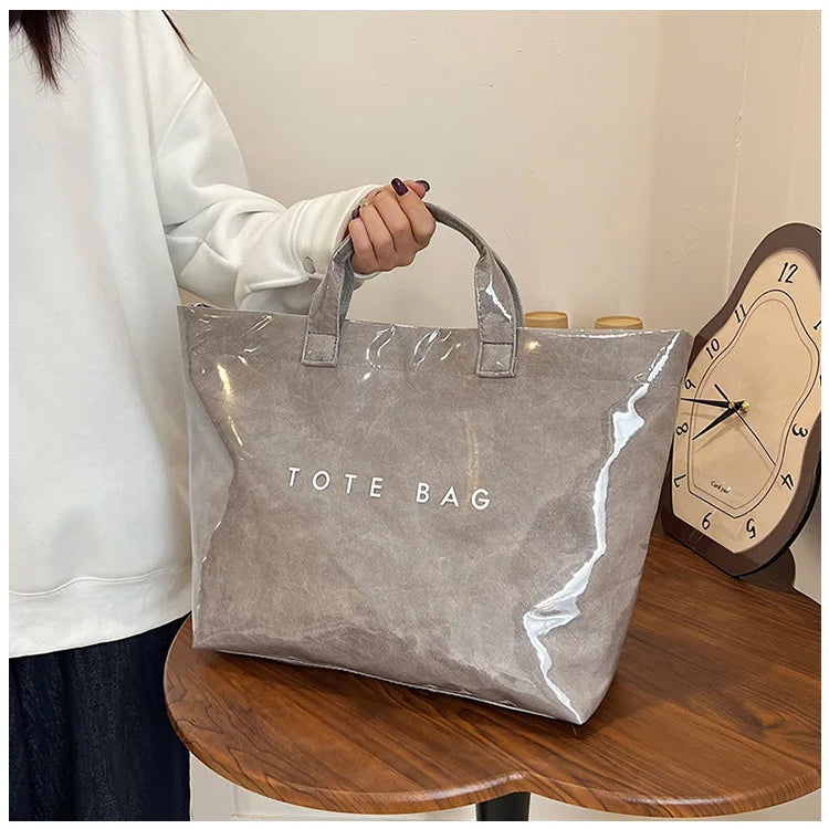 A light gray transparent waterproof PVC tote bag is displayed on a wooden table next to a person's arm.