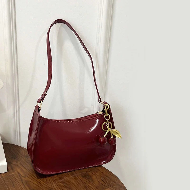 A maroon PU leather shoulder bag with a gold cherry pendant hanging against a white wall.