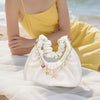 A white elegant metal bag with gold charms and keychains resting on a white fabric near the ocean.