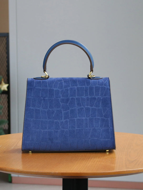 A deep blue crocodile pattern tote bag with a leather handle resting on a wooden table.