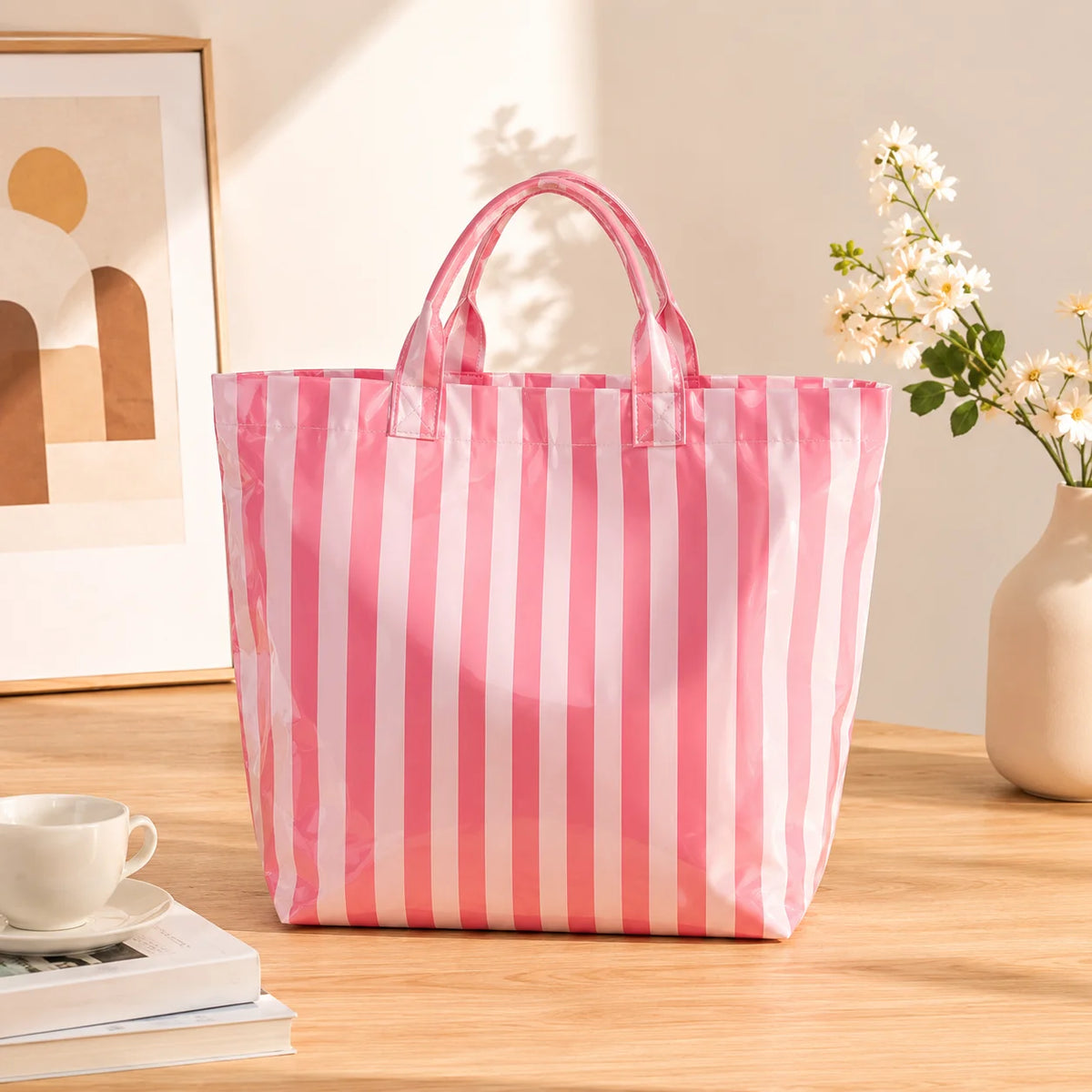 A pink and white striped waterproof PVC shoulder tote bag resting on a wooden table next to a cup and some books.