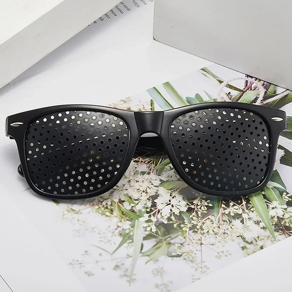 A pair of black precision vision correction glasses resting on a white surface with greenery.