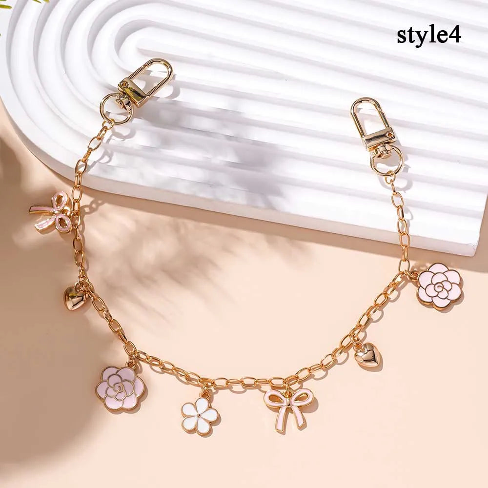 Accesorio de bolso con cadena tono dorado, adornado con charms de corazón y flor de cerezo sobre fondo blanco.