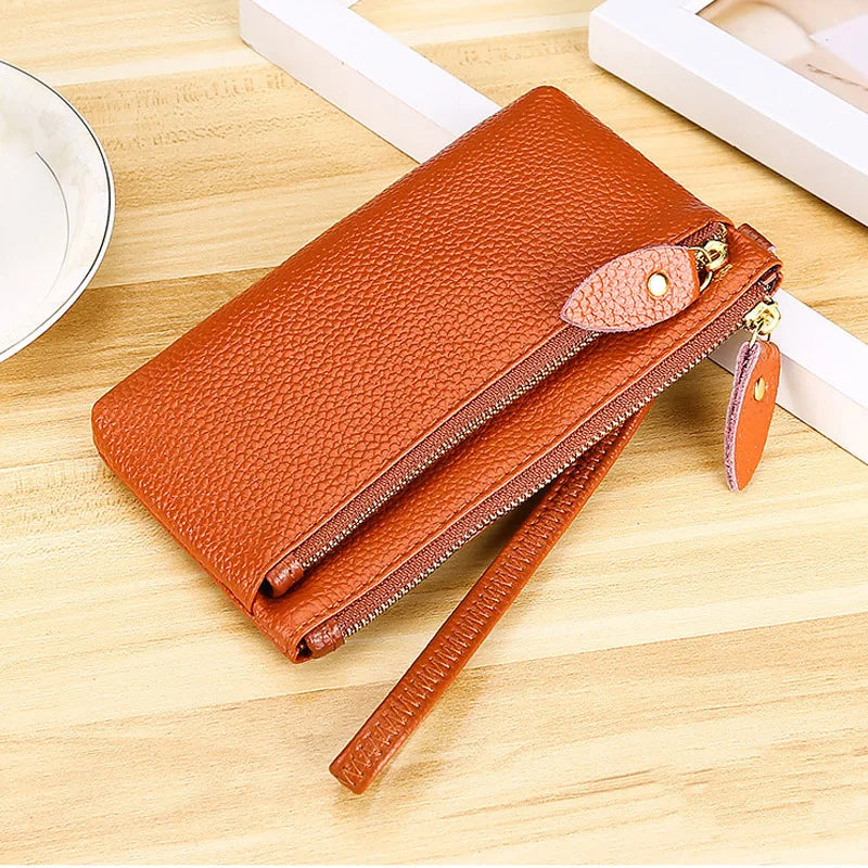 A brown genuine leather wristlet clutch wallet with a zipper closure resting on a wooden surface.