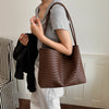 A person holds a brown crocodile-textured PU bucket shoulder bag against a gray t-shirt.