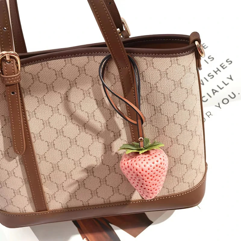 Un sac à main en cuir marron avec un charm de fraise rose attaché à la sangle.