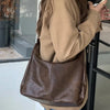 A person wearing a beige jacket and blue jeans holds a brown leather tote bag with a crossbody strap.