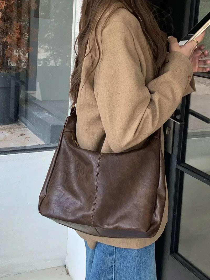 A person wearing a beige jacket and blue jeans holds a brown leather tote bag with a crossbody strap.