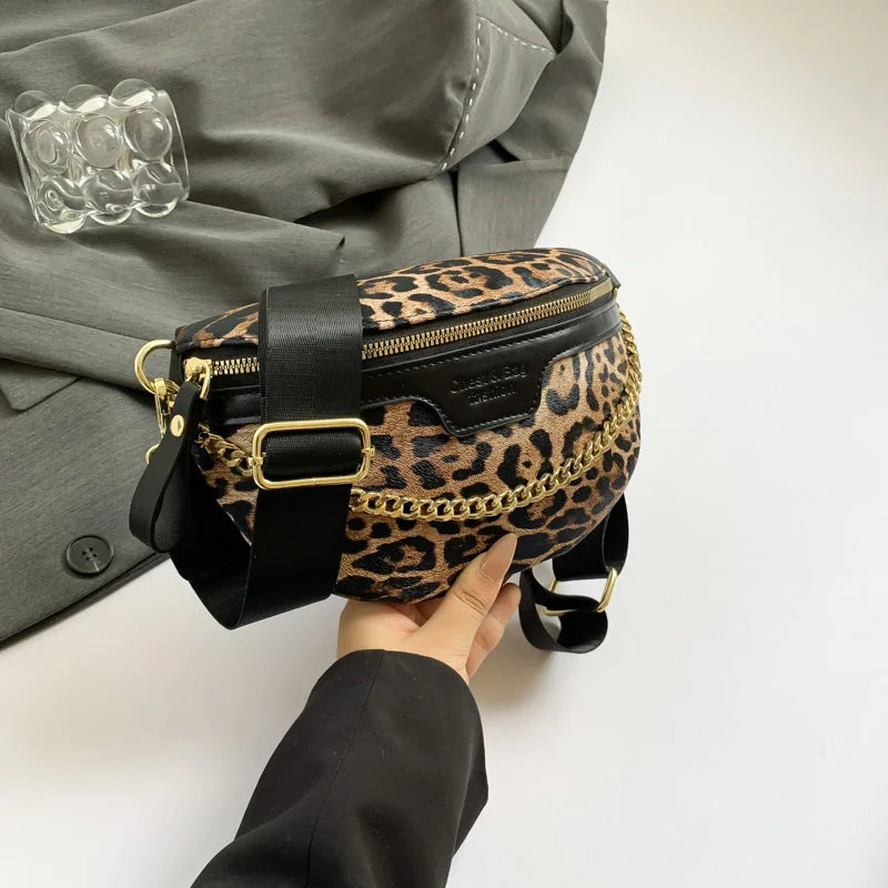 Eine Hand hält eine schwarz-braune Leopardenmuster-Crossbody-Tasche mit goldfarbenem Zierwerk vor einem grauen Blazer.