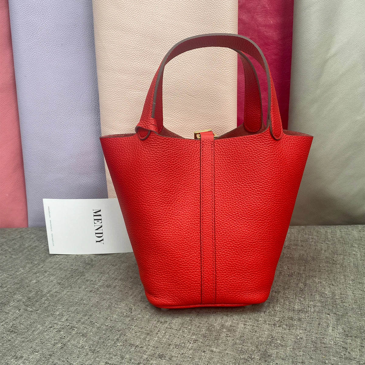 A bright red leather bucket bag with a top handle displayed against a backdrop of pastel colored fabric.