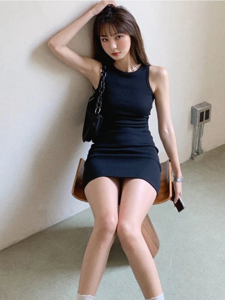 A woman wearing a black sleeveless bodycon tank dress with a slim fit mini style, posing against a light wall.