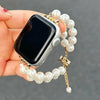 Eine Hand hält ein iPhone mit einem Perlenarmband, das goldfarbene Glieder und weiße Kristallperlen aufweist.