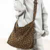 A brown leopard print canvas shoulder bag with an adjustable strap is shown against a person wearing a gray hoodie.
