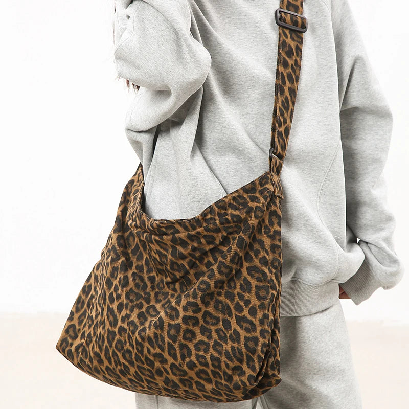 A brown leopard print canvas shoulder bag with an adjustable strap is shown against a person wearing a gray hoodie.