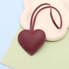 A maroon leather heart-shaped keychain charm is shown resting on a light green card next to a pale pink background.