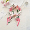 Foulard floral de soie rose et vert noué en nœud, posé sur un tissu blanc à côté d'un flacon de parfum.