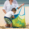 A woman is handing a bright green mesh storage bag to a young boy on a sandy beach.