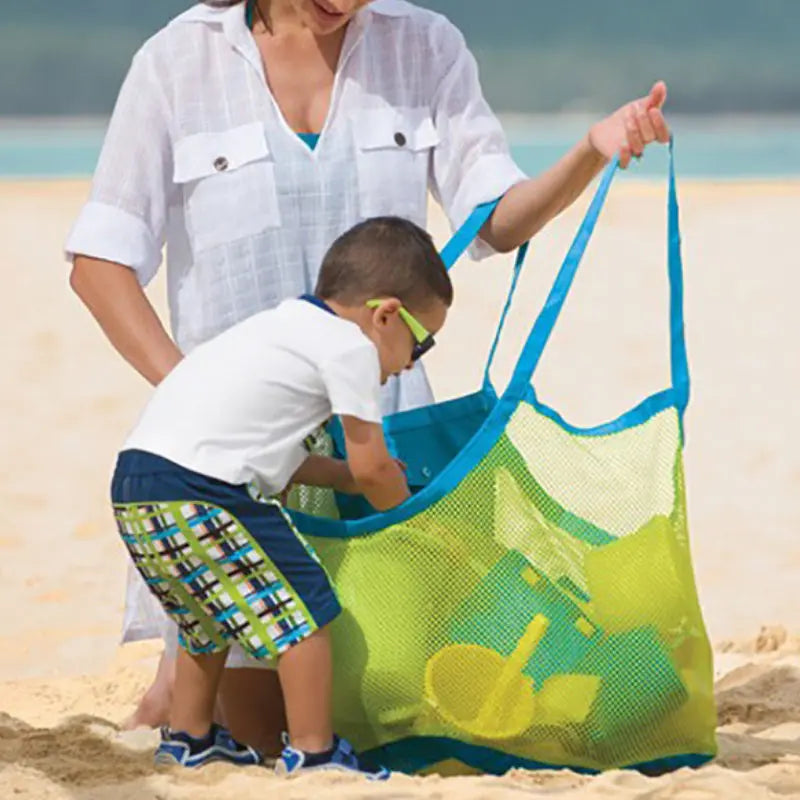 A woman is handing a bright green mesh storage bag to a young boy on a sandy beach.