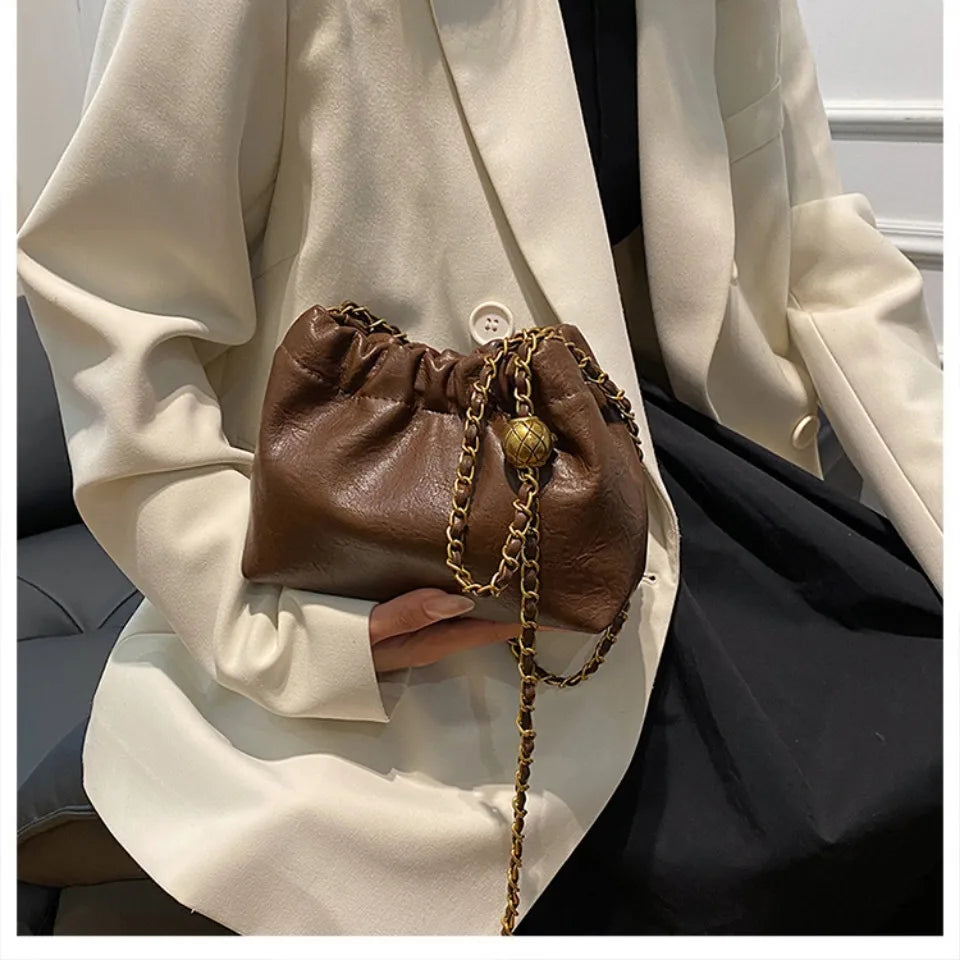 A brown PU leather chain shoulder bag is held against a cream blazer and black dress.