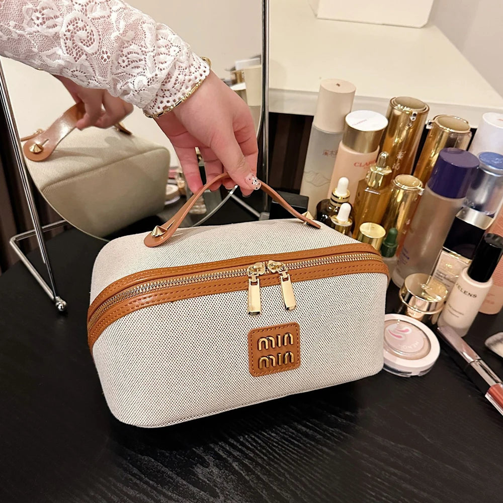 A beige travel toiletry bag with a brown leather trim and gold hardware is shown on a dark surface.