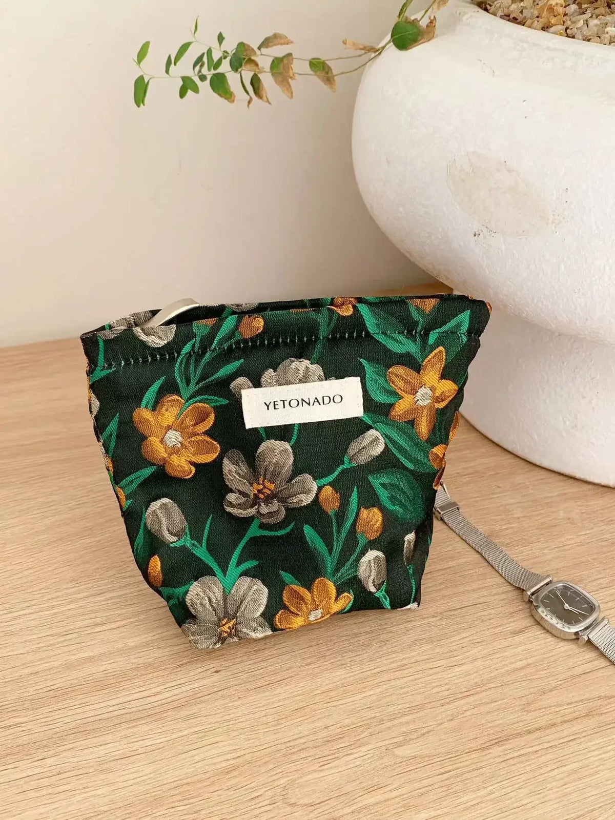 A dark green cosmetic pouch featuring a floral pattern of yellow and gray flowers resting on a wooden surface.