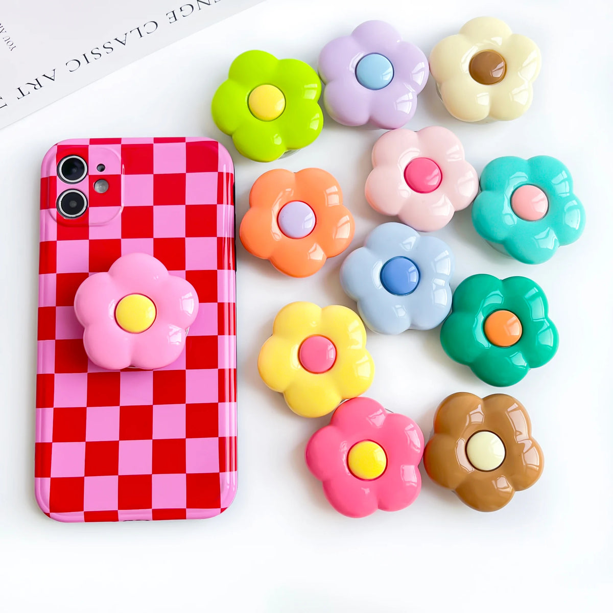 A pink checkered phone case is displayed next to several colorful, flower-shaped silicone grips.