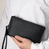 A hand holds a black textured clutch bag with a zipper and strap against a white shirt.