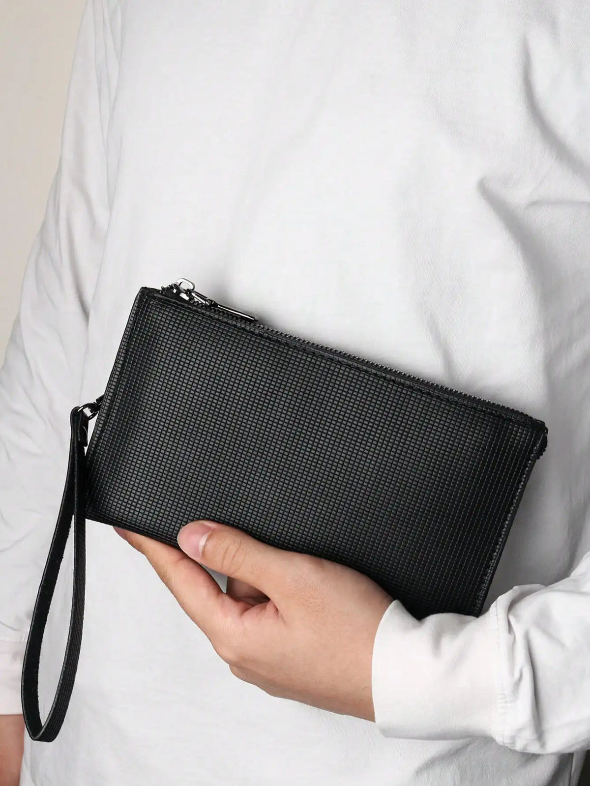 A hand holds a black textured clutch bag with a zipper and strap against a white shirt.