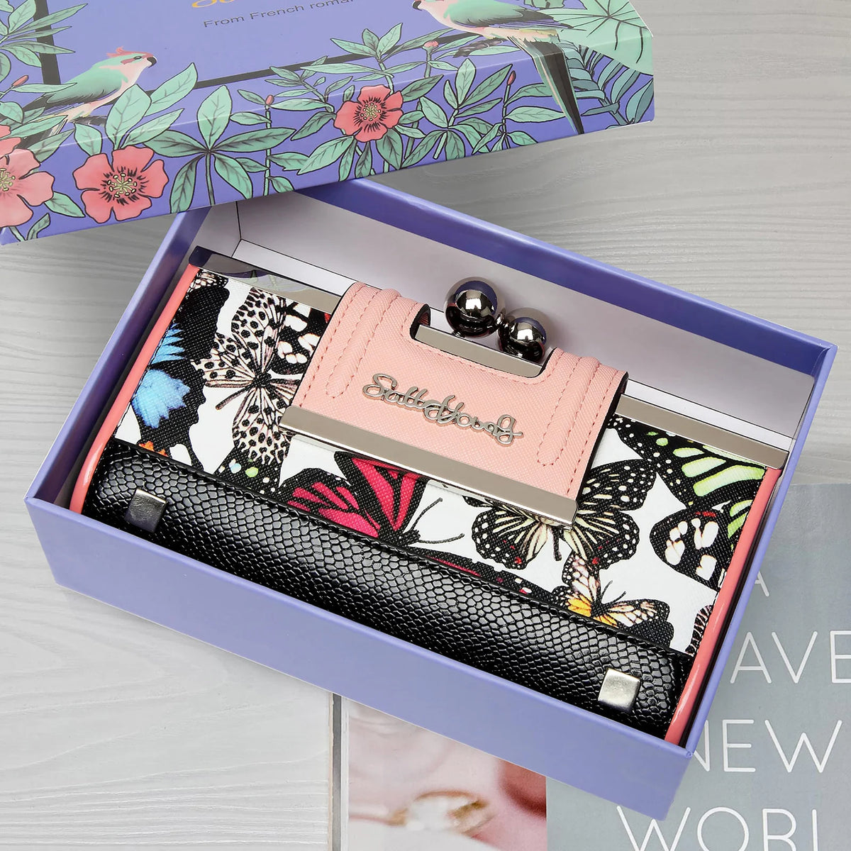 Cartera de cuero rosa con estampado floral de mariposa, exhibida dentro de una caja morada.