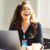 A woman smiling while holding a retractable badge reel with a colorful lanyard and a laptop.