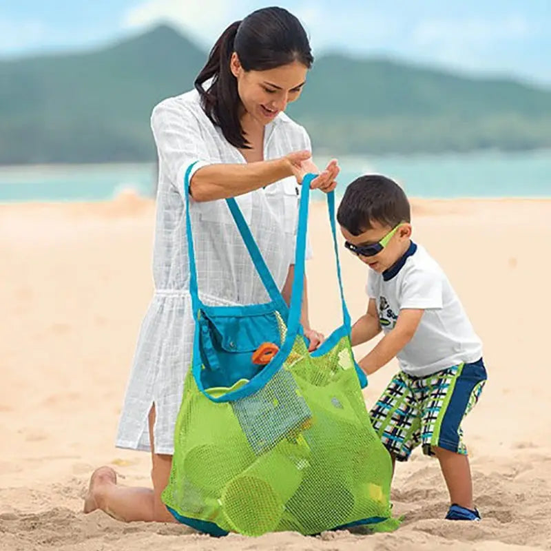 A woman is using a multi-use mesh beach and storage bag for toys with a young child on a sandy beach.