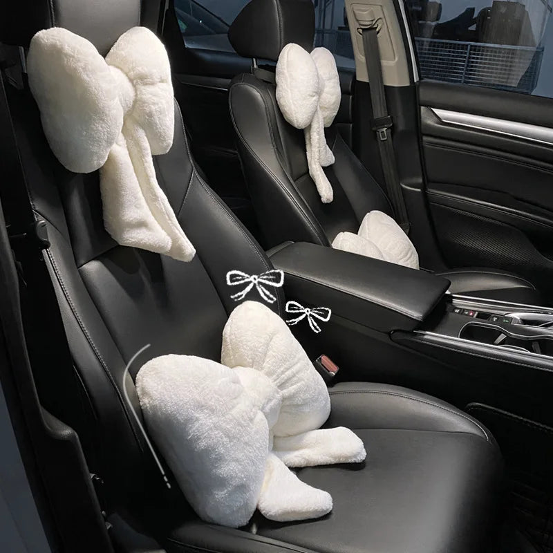A black car interior showing a seat with a white fluffy bowknot headrest pillow and lumbar support cushion.