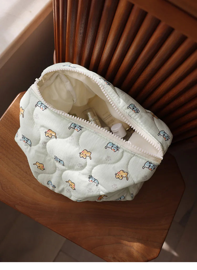A white quilted cosmetic storage pouch with a light blue and orange cat print is resting on a wooden surface.