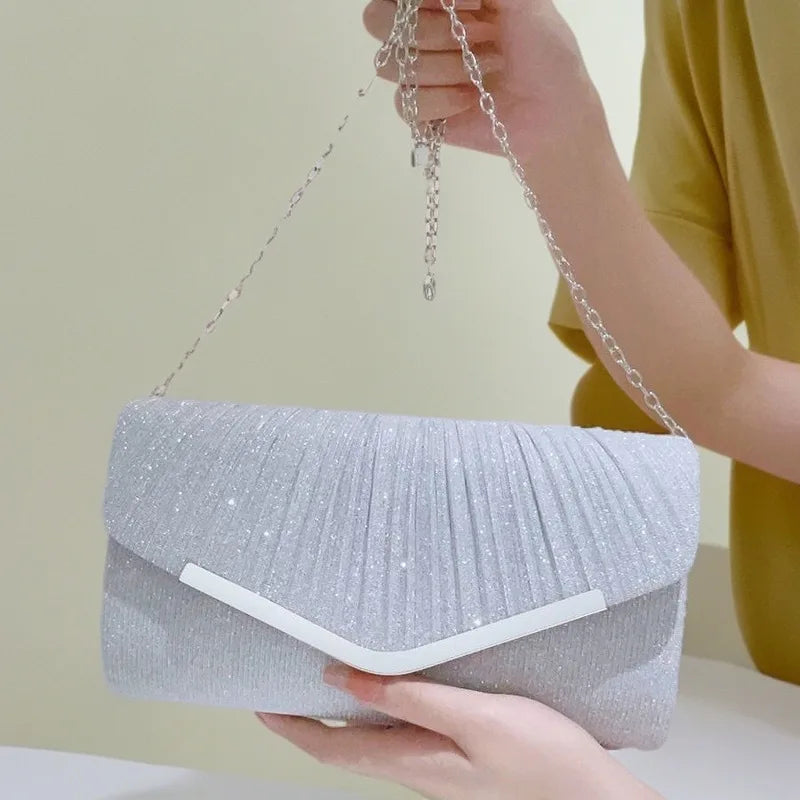 A hand holds a silver glitter pleated crossbody clutch bag with a detachable chain against a light background.