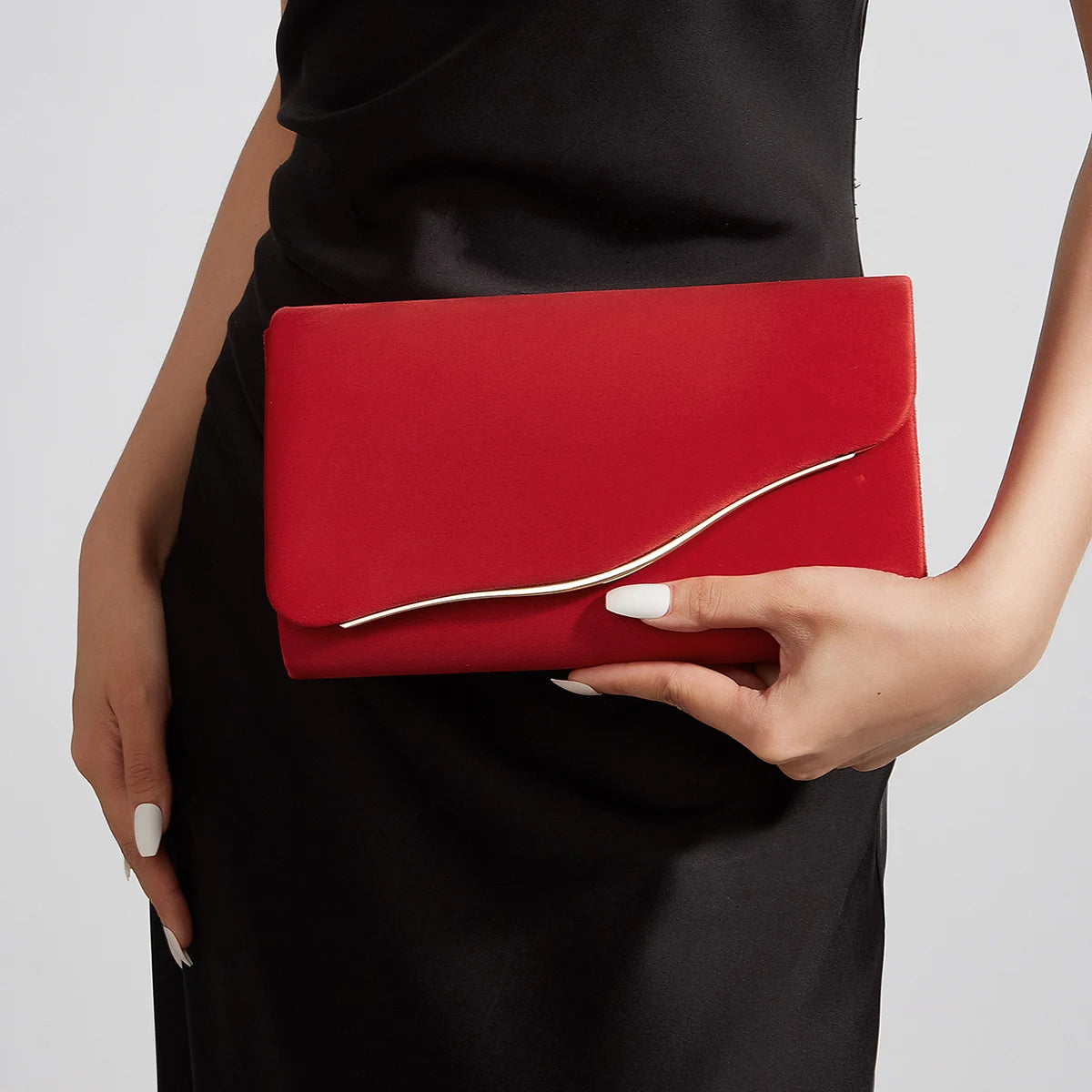 A hand holds a red velvet evening clutch with a silver detachable shoulder strap against a black dress.