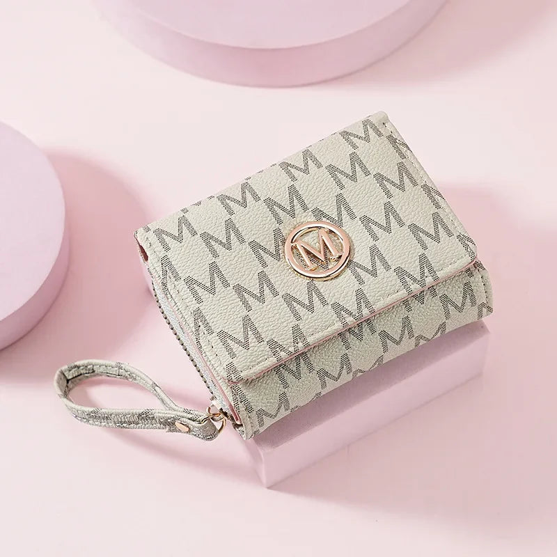 A light green letter zipper wristlet wallet with a gold monogram is displayed on a pink surface.