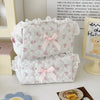 A white cosmetic storage pouch with pink floral embroidery and a pink bow, displayed next to a framed piece of art.