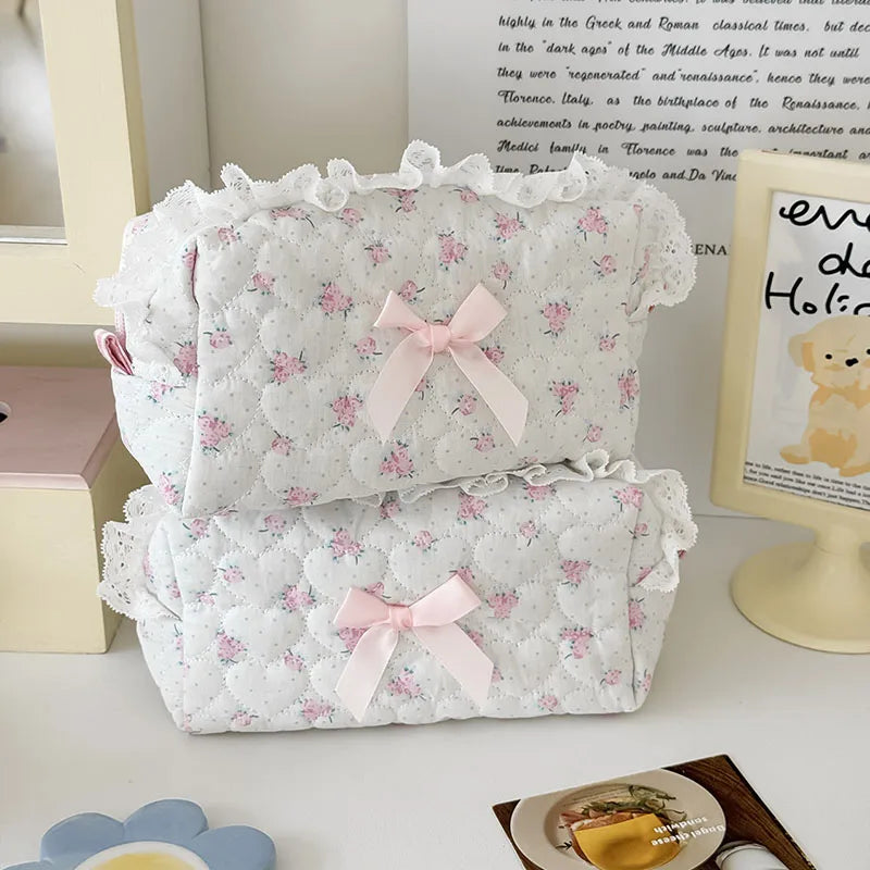 A white cosmetic storage pouch with pink floral embroidery and a pink bow, displayed next to a framed piece of art.