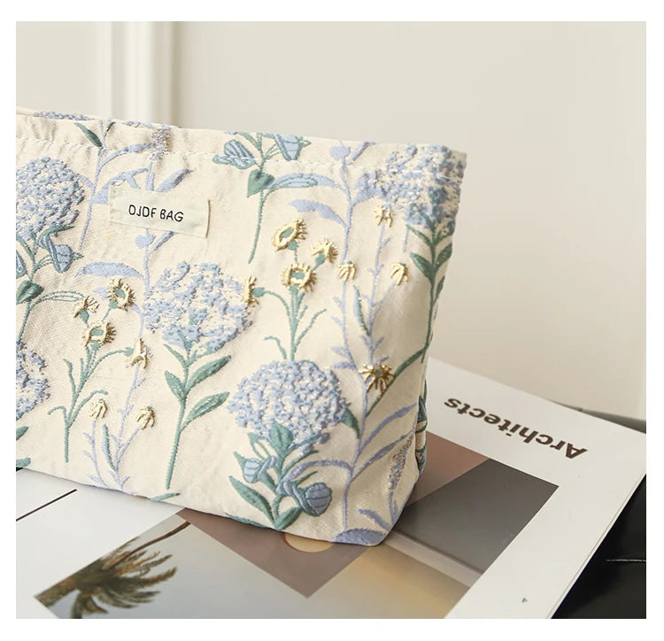 A floral canvas makeup bag featuring blue and green botanical embroidery resting on a white surface.
