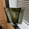 A dark olive green leather shoulder bag with a wide strap featuring colorful embroidery hanging in front of a window.