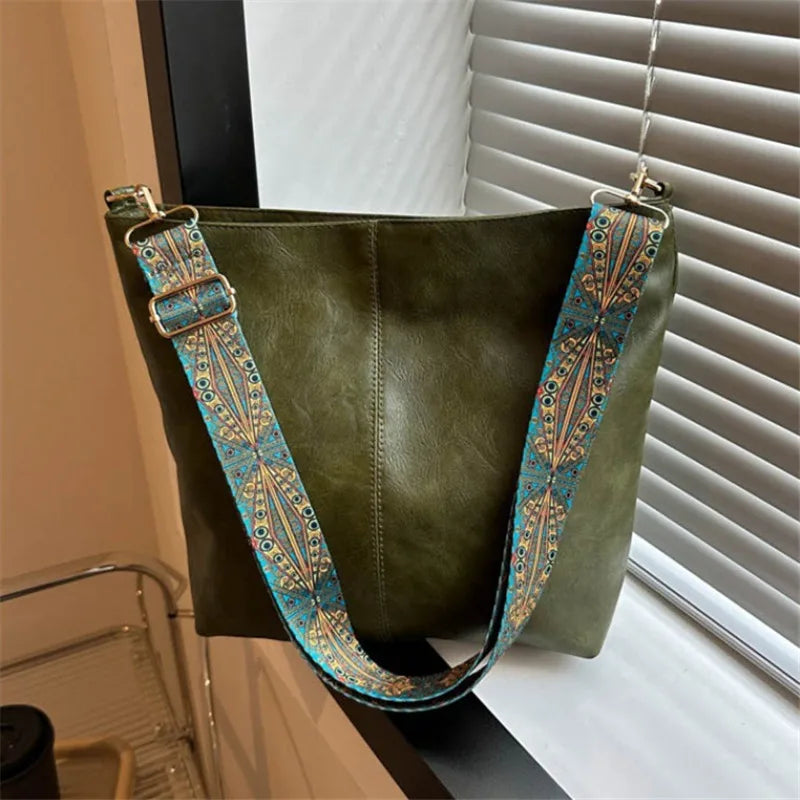 A dark olive green leather shoulder bag with a wide strap featuring colorful embroidery hanging in front of a window.