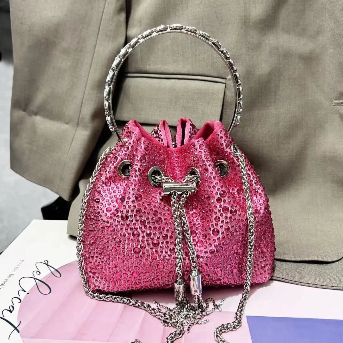 A pink crystal-embellished evening clutch with a silver chain is displayed against a light gray suit jacket.
