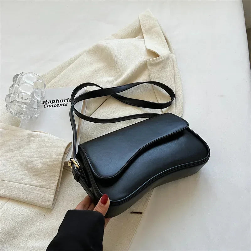 Un bolso cruz de cuero negro con una correa delgada junto a una chaqueta color crema sobre una superficie blanca.