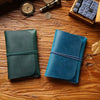 A dark teal leather passport holder and a blue leather card wallet resting on a wooden surface with a vintage clock and loose