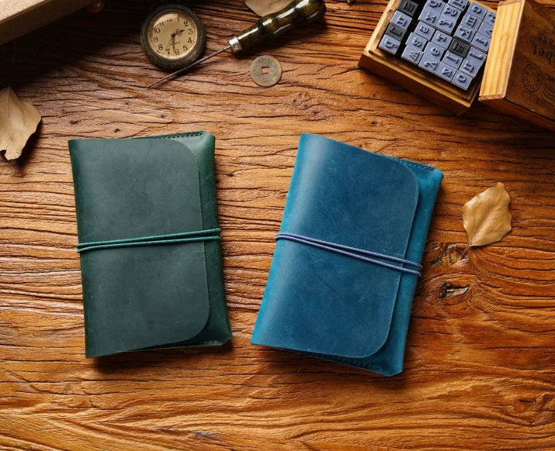 A dark teal leather passport holder and a blue leather card wallet resting on a wooden surface with a vintage clock and loose